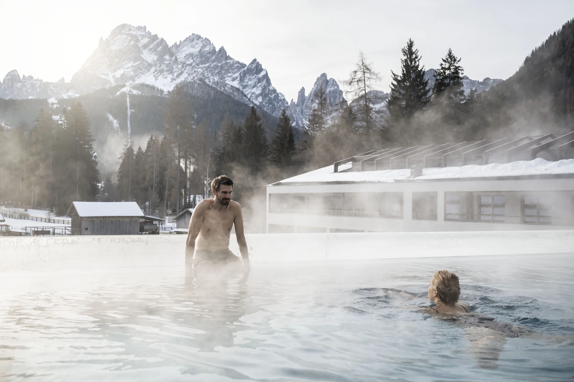 Zwei Personen im Außenpool mit dampfendem Wasser vor schneebedeckten Bergen