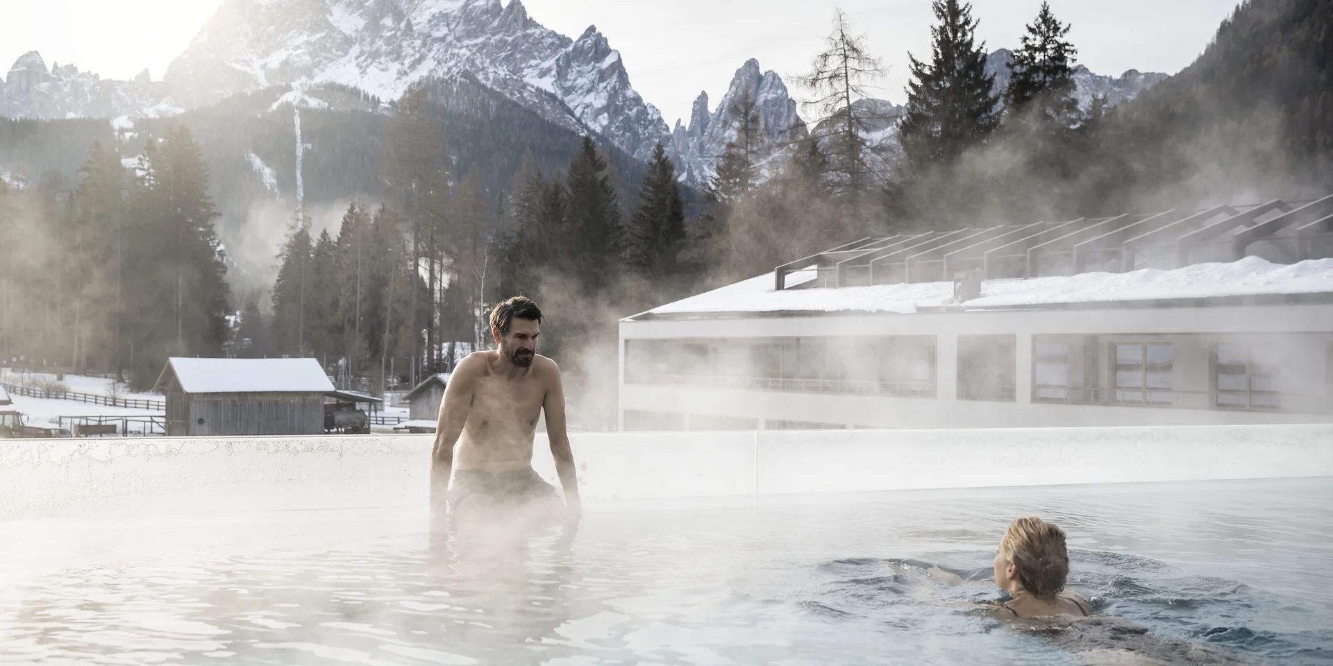 Due persone in piscina all'aperto con acqua fumante davanti a montagne innevate