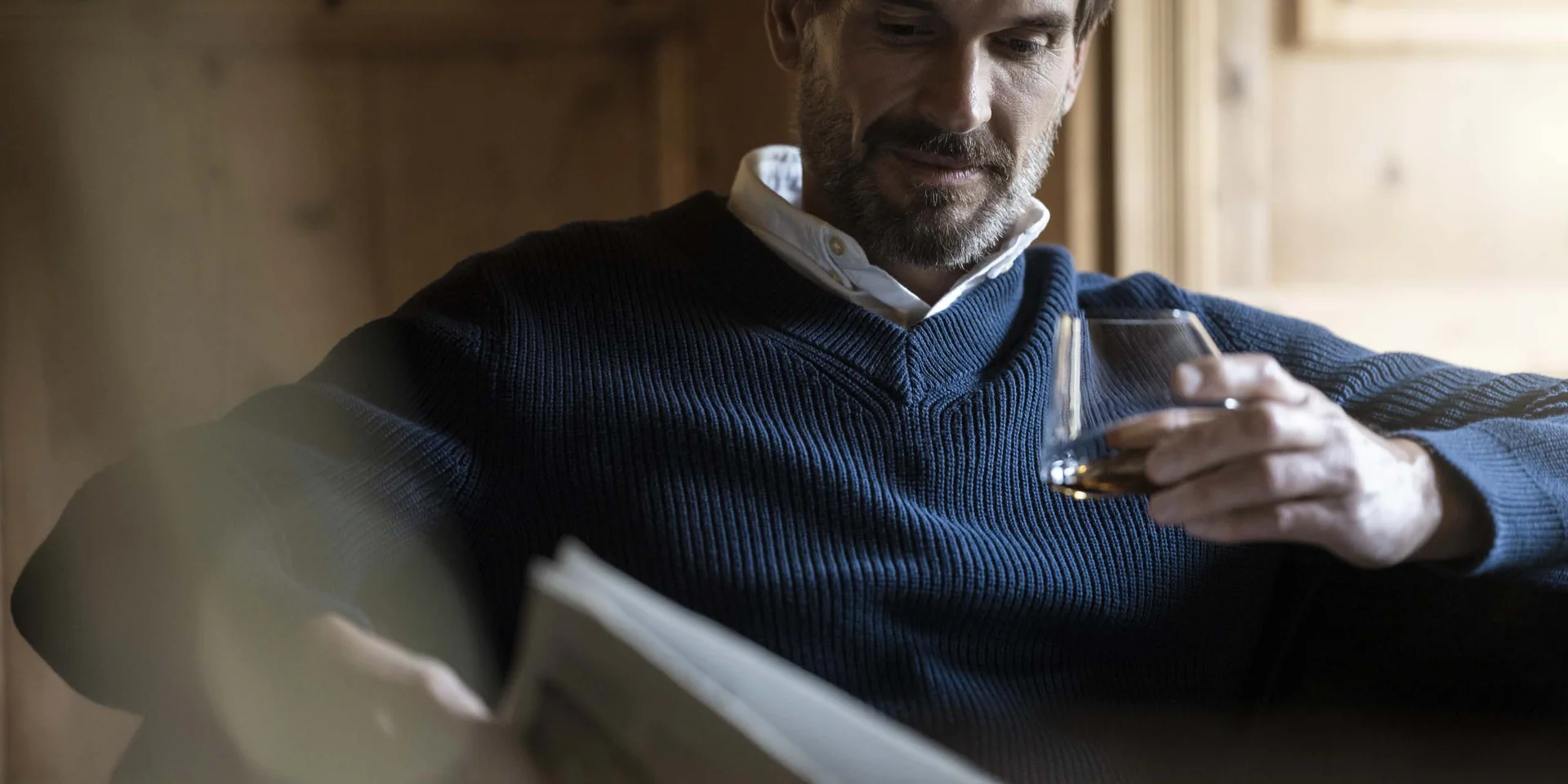 Mann in blauem Pullover liest Zeitung und hält ein Glas Whisky