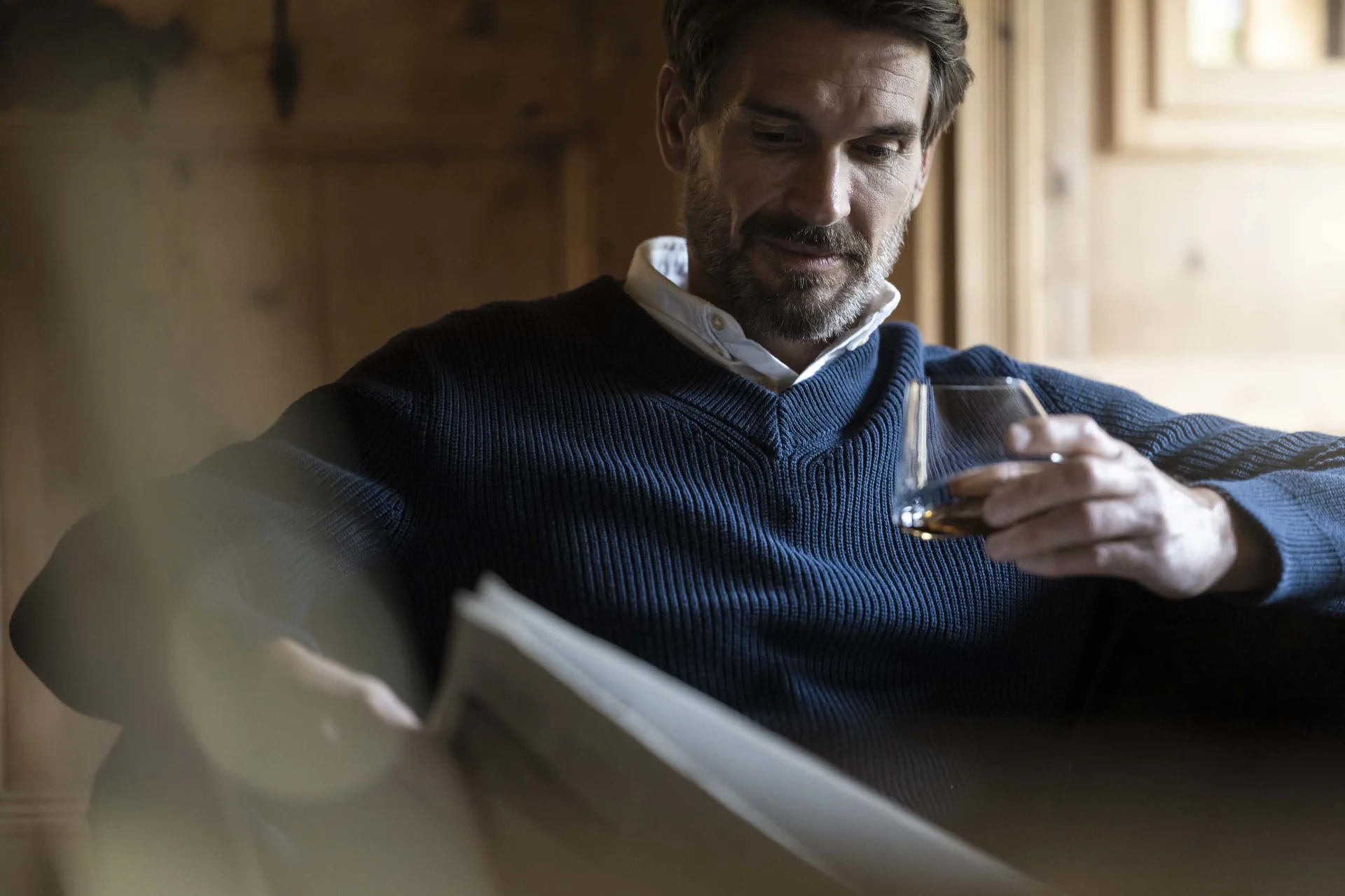 Mann in blauem Pullover liest Zeitung und hält ein Glas Whisky