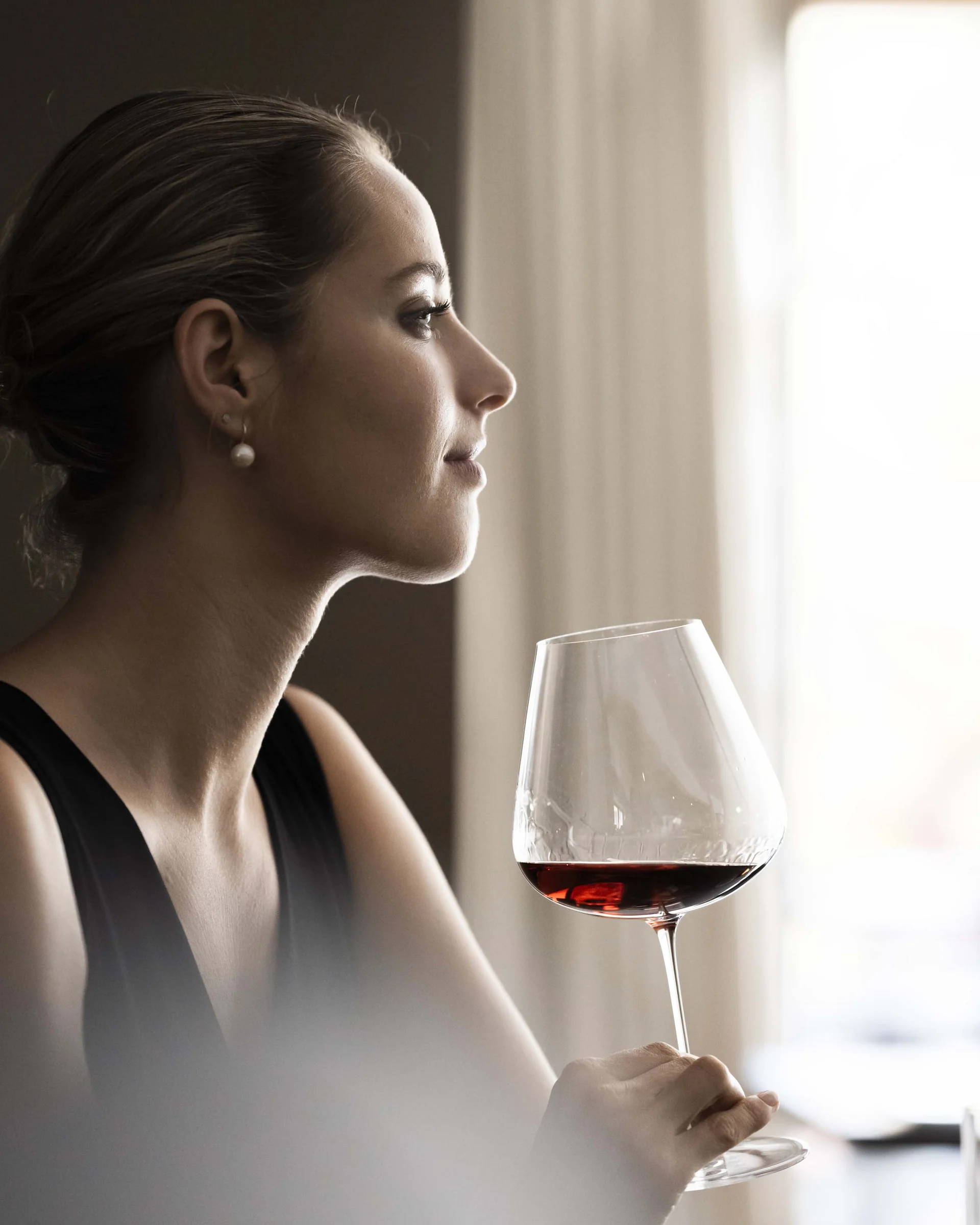 Frau in elegantem Kleid hält Weinglas mit Rotwein und blickt aus Fenster