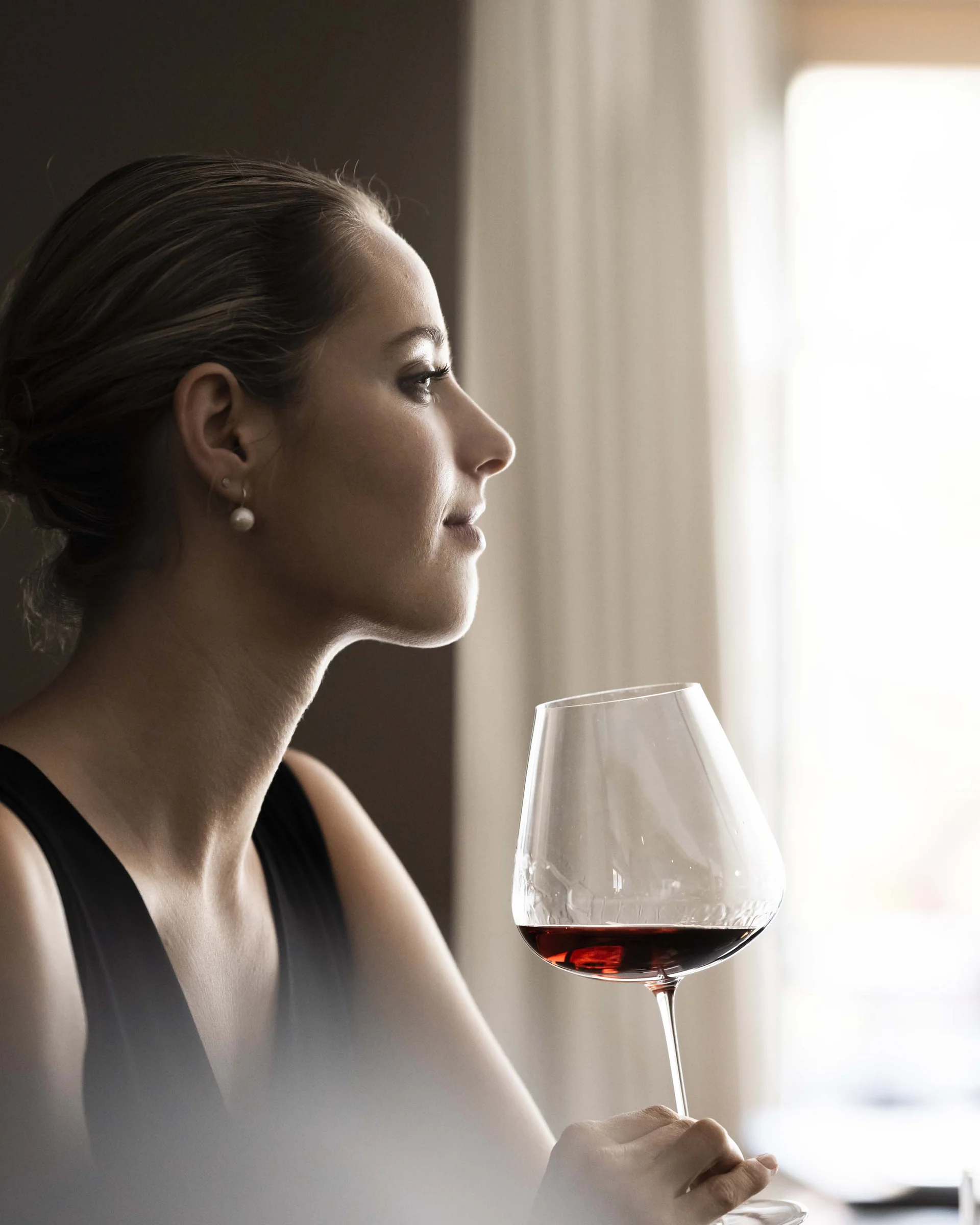 Frau in elegantem Kleid hält Weinglas mit Rotwein und blickt aus Fenster