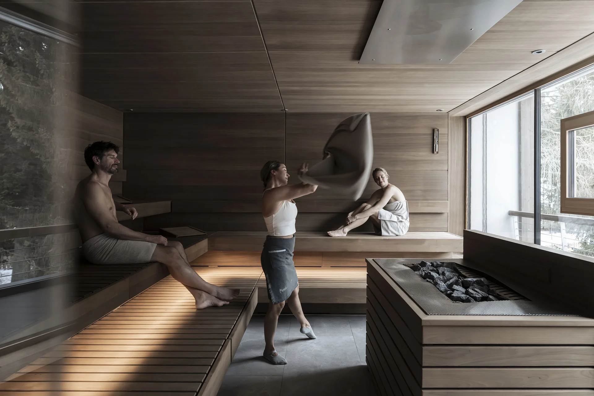 Drei Personen entspannen sich in einer Sauna mit großen Fenstern.