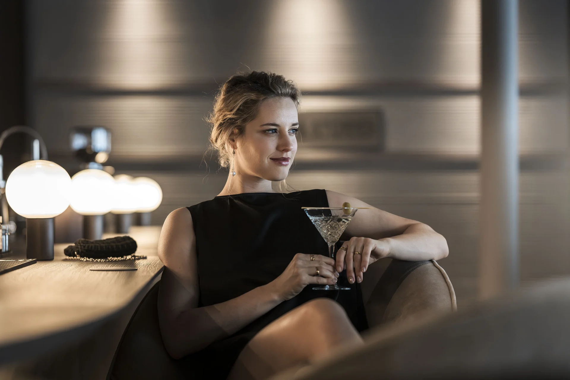 Frau in schwarzem Kleid sitzt an Bar mit Cocktailglas und Oliven