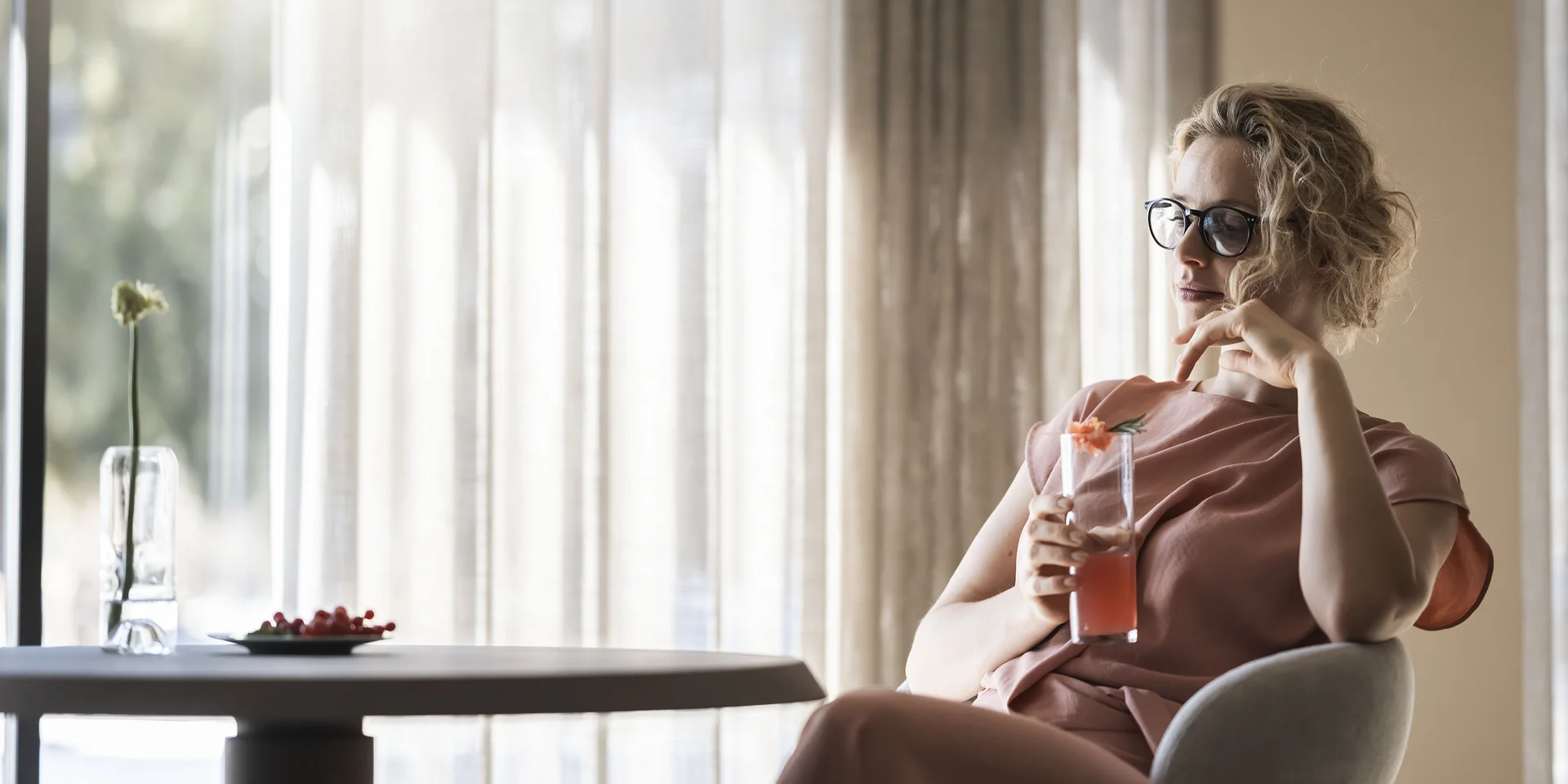 Donna con occhiali seduta da sola che beve un succo in una stanza luminosa