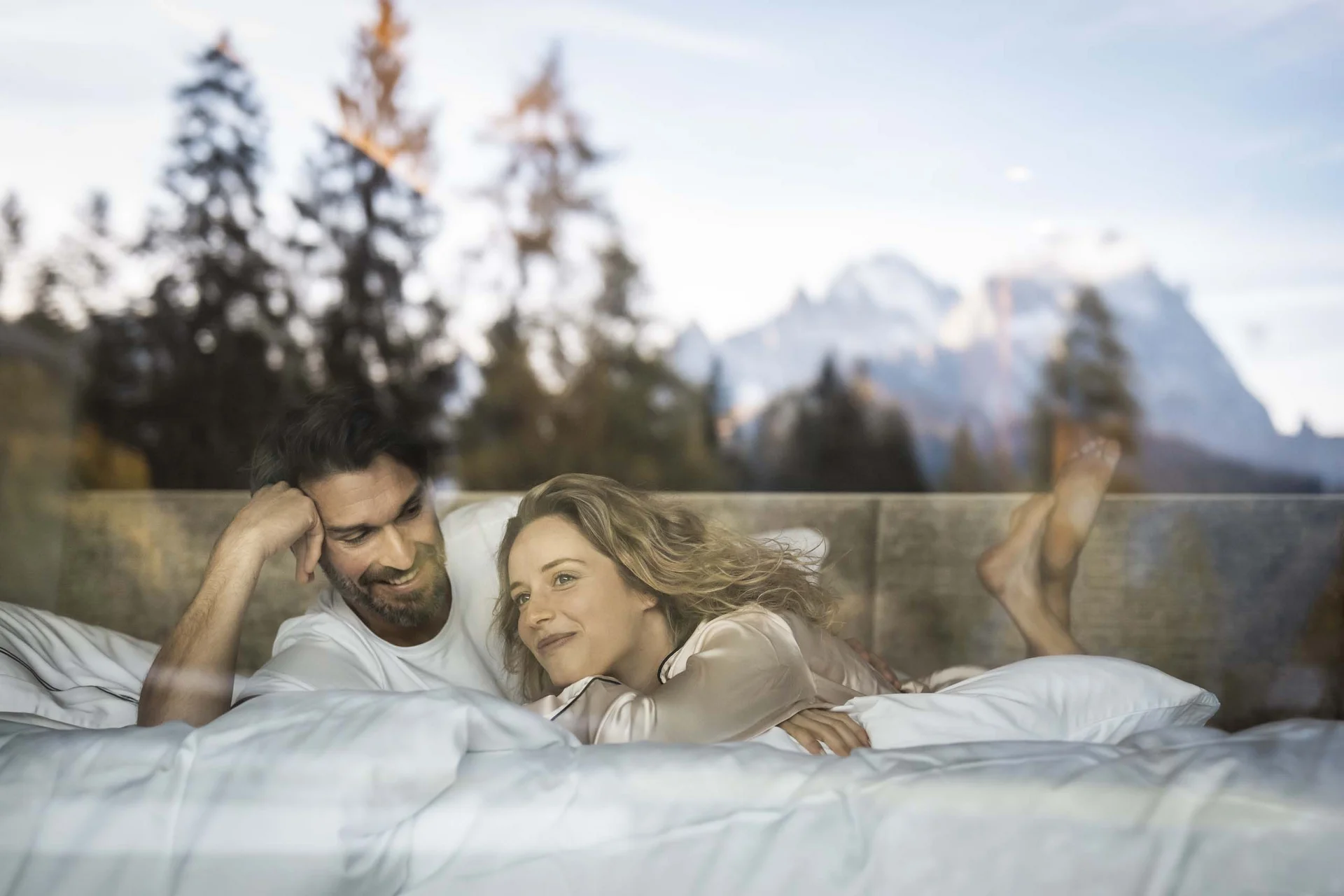 Paar liegt entspannend im Bett mit Bergblick und Bäumen draußen