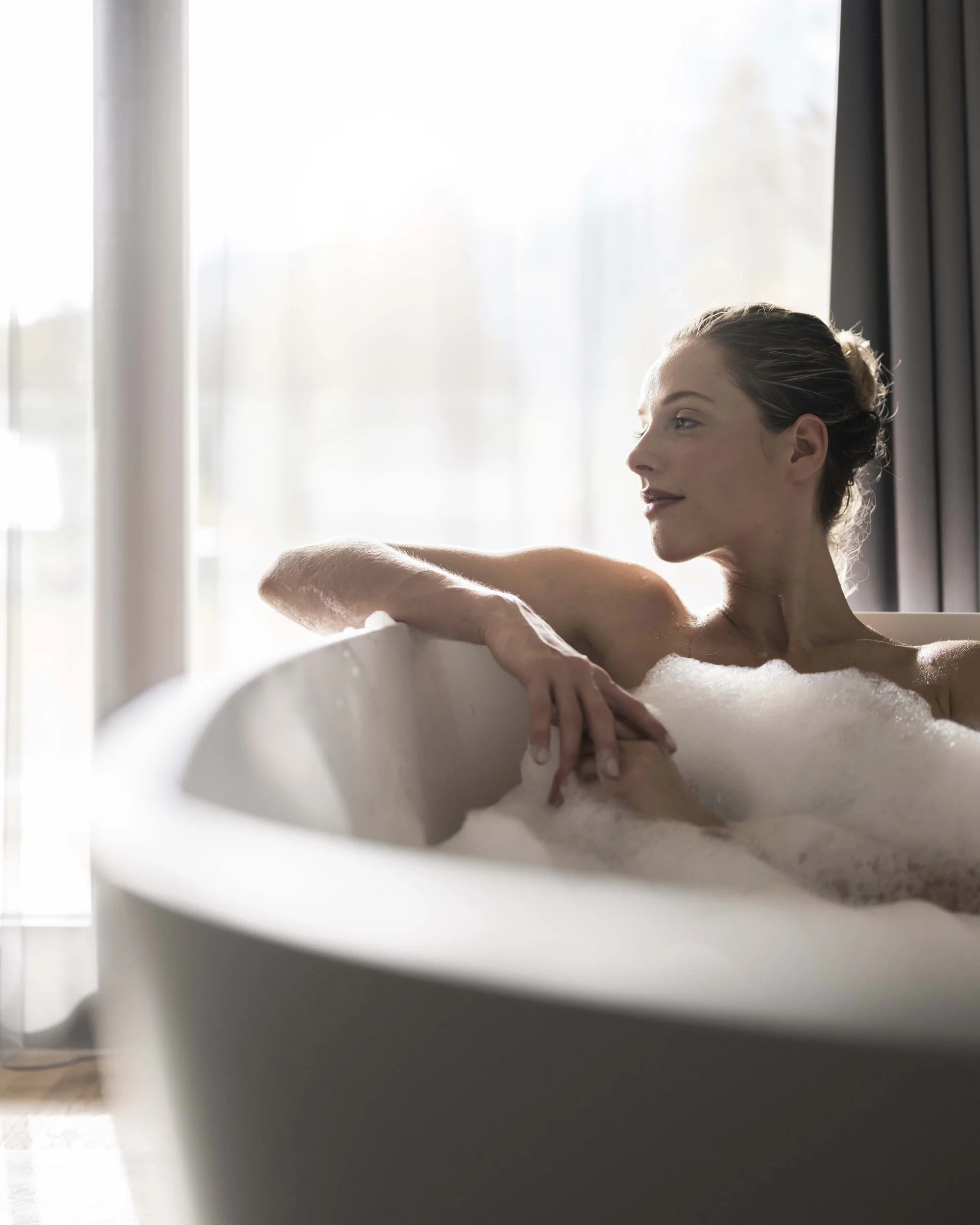 Frau entspannt in Badewanne mit Schaum vor hellem Fenster