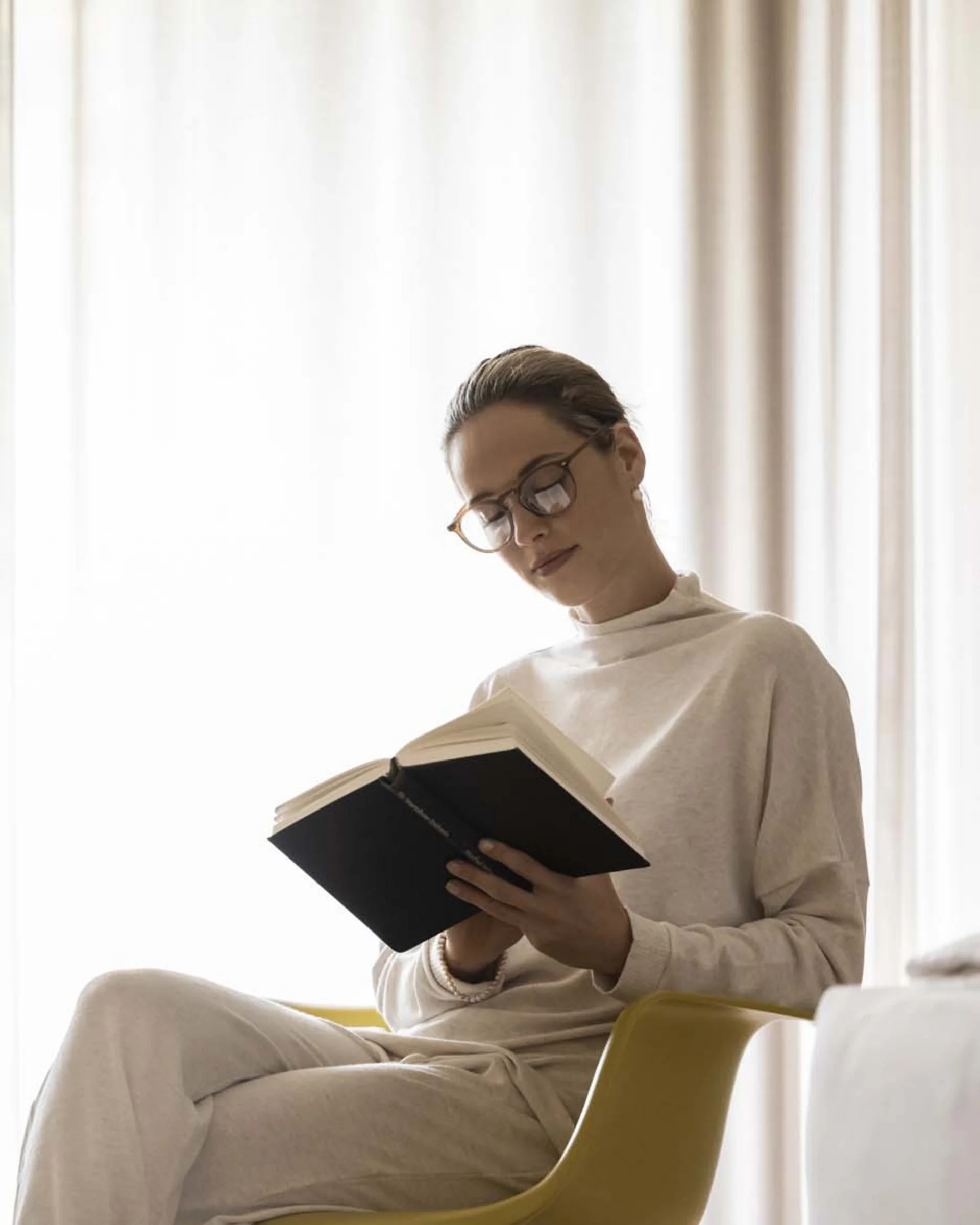 Frau liest Buch entspannt auf einem gelben Schaukelstuhl im Wohnzimmer